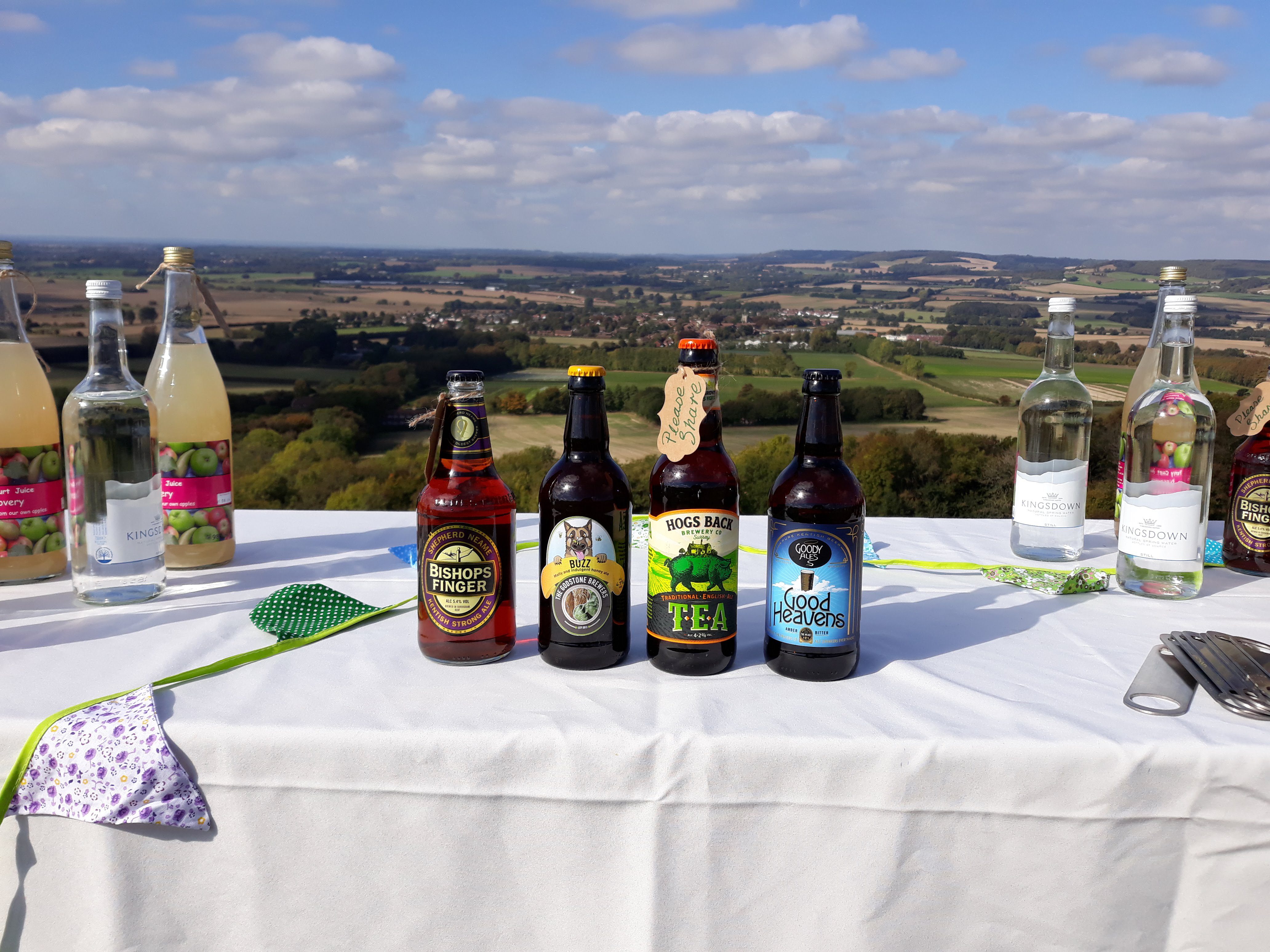 View across grass and crop fields, with picnic table and drinks in the foreground.