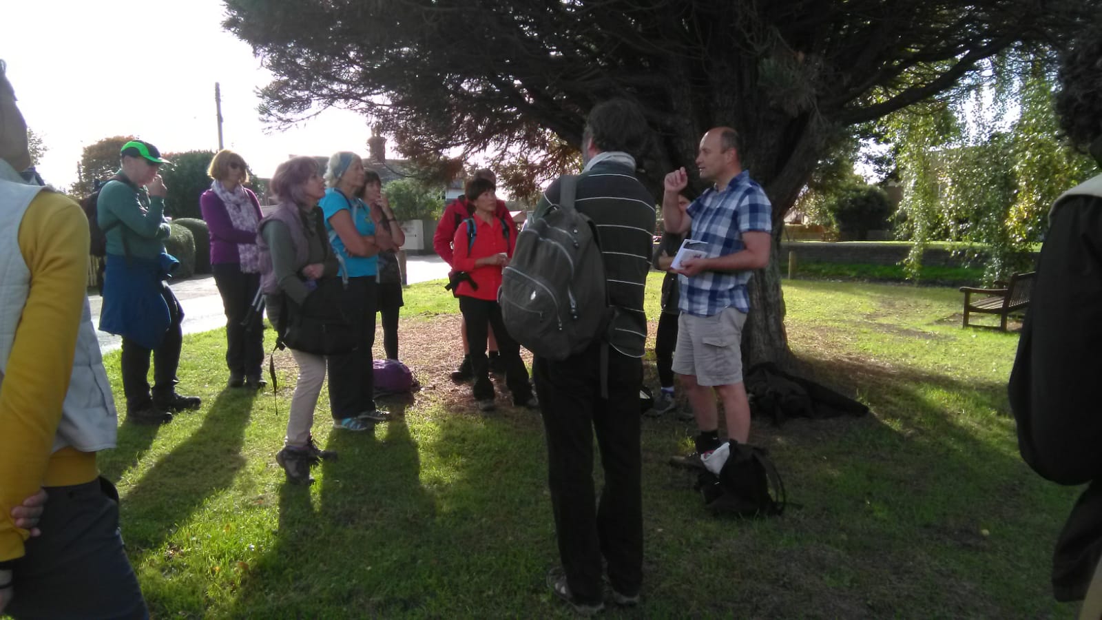 Group of people standing, looking at tour guide, under a trees on grass.