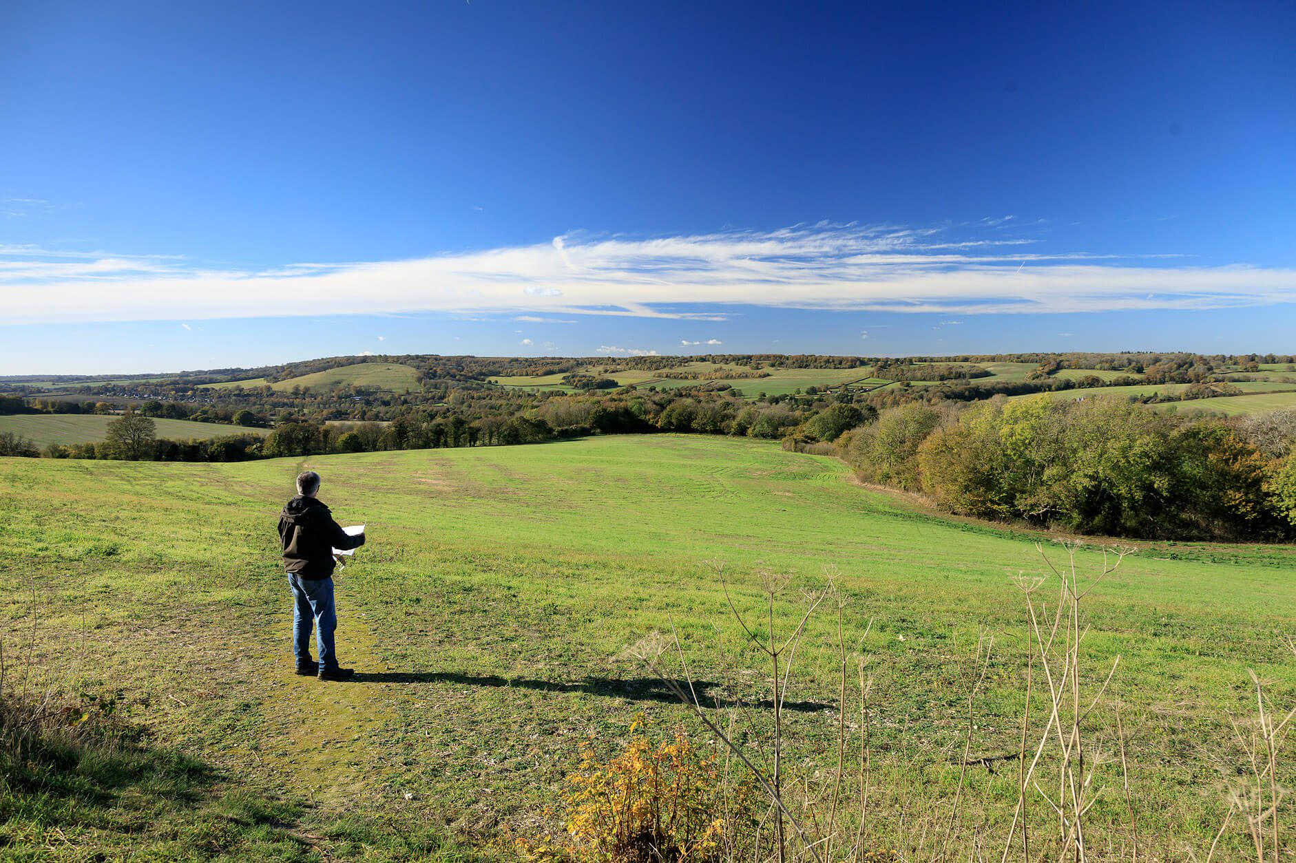 Green fields and trees in the distance, views across Stour Valley. Blue sky. Person looking at view with map in hand.