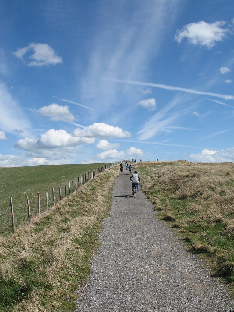 Cyclists on path, heading away from camera. Grass fields on both sides of path and blue skies.