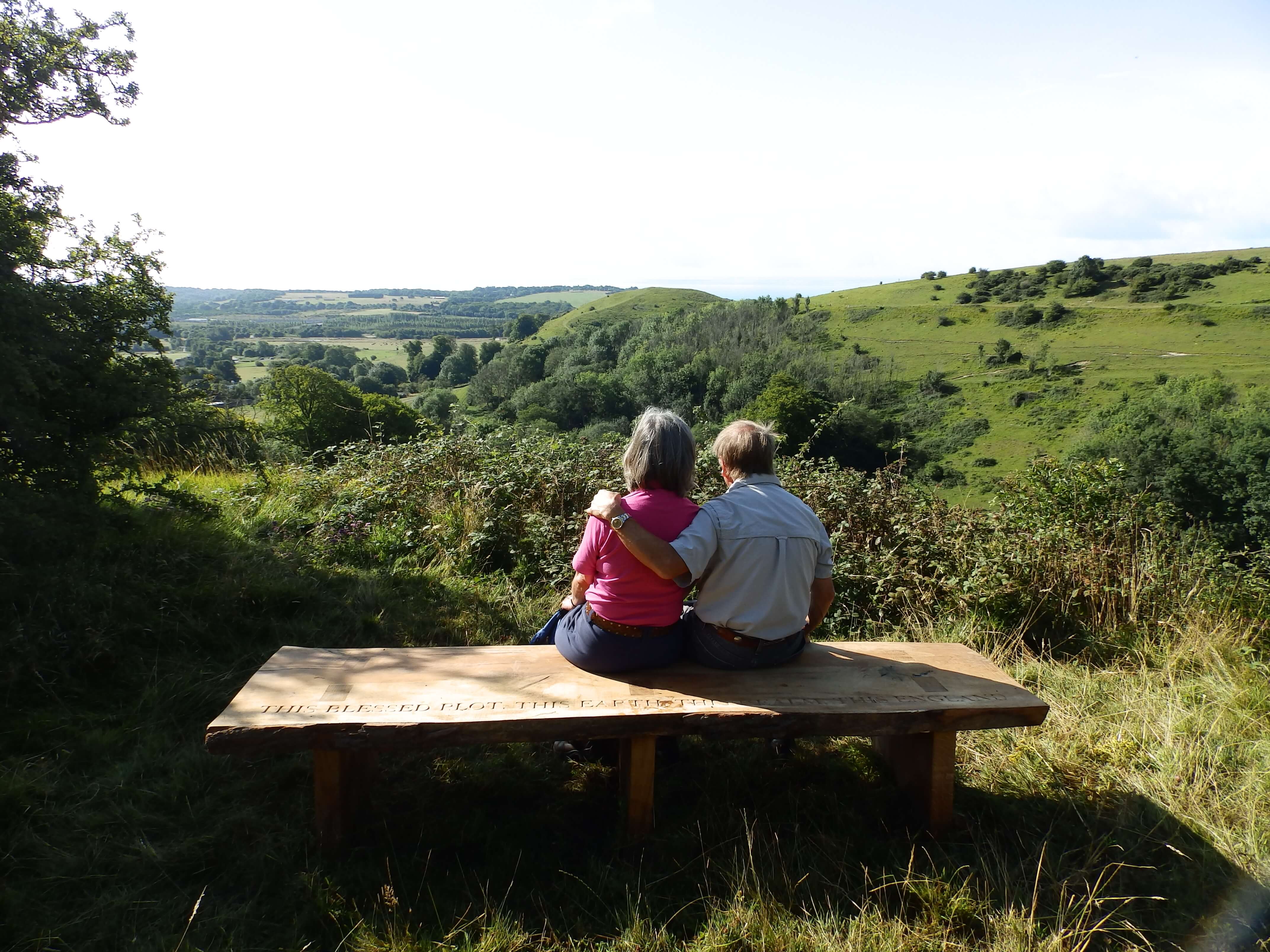 Two people seat on bench overlooking view across green fields and trees.