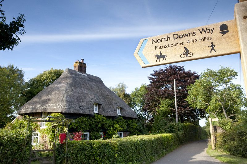 North Downs Way sign overlooking a thatched roof house on a country road, sunny day.