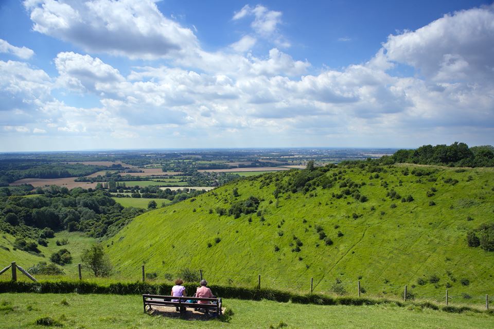 Views across grass fields and trees, with two people sitting at bench in foreground.