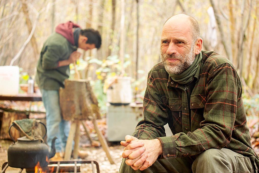 Person in woodland, sitting near pot on campfire, with another person in background working on a tree stump.