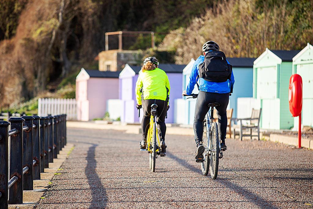 Two people cycling along sea front, with beach huts on the right. Cycling away from camera.