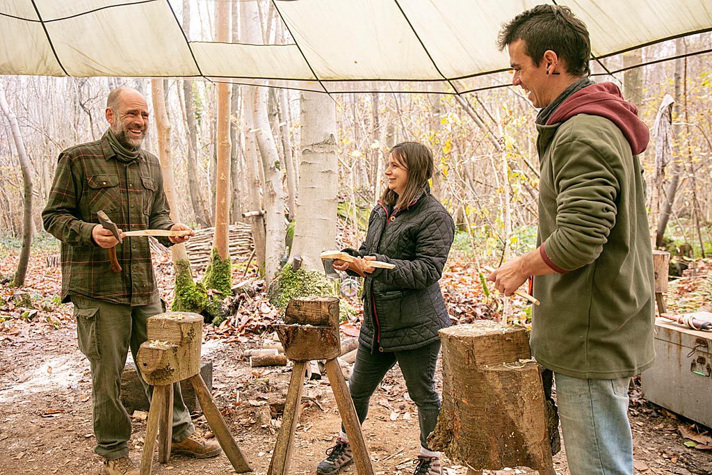 Three people working on making wooden spoons, in a woodland clearing, under a canopy.