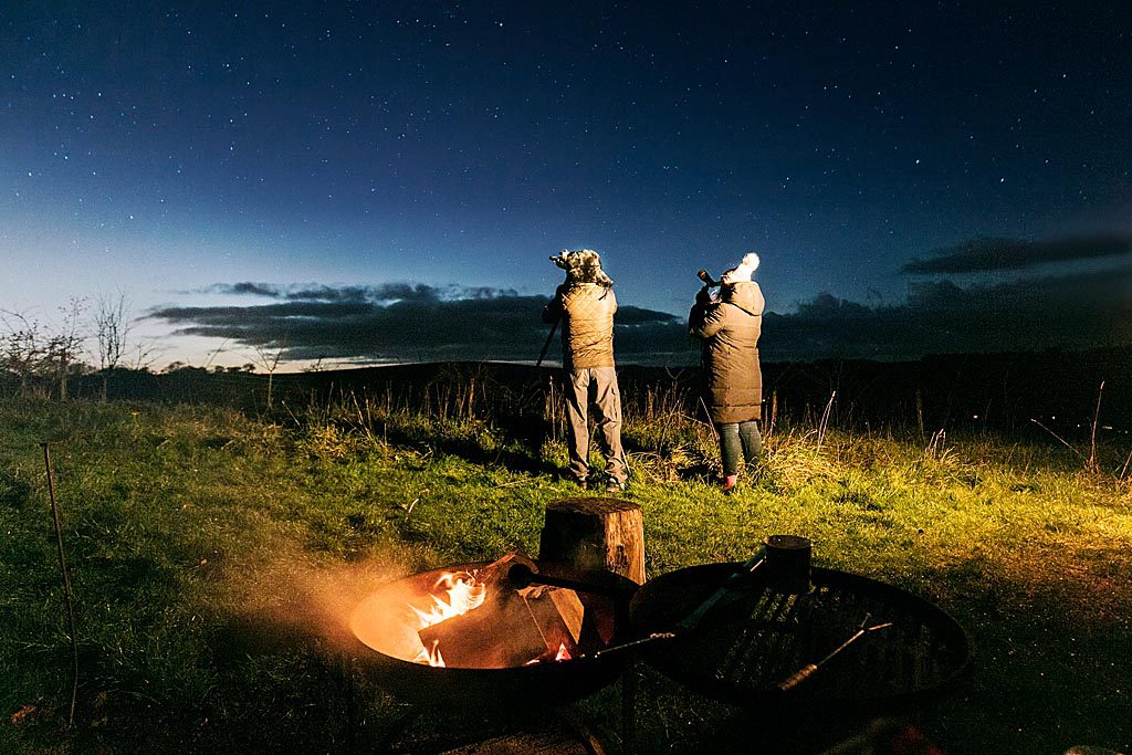 Two people standing looking at the night sky, through a telescope, in a field with camp fire behind them.