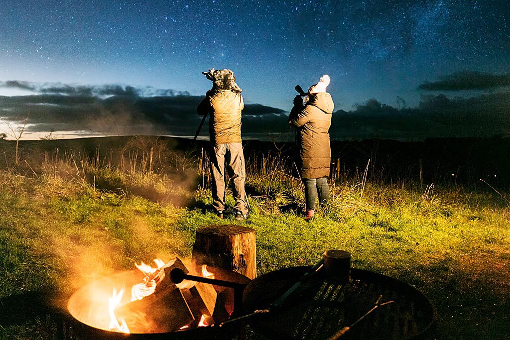 Two people standing looking at the night sky, through a telescope, in a field with campfire behind them.