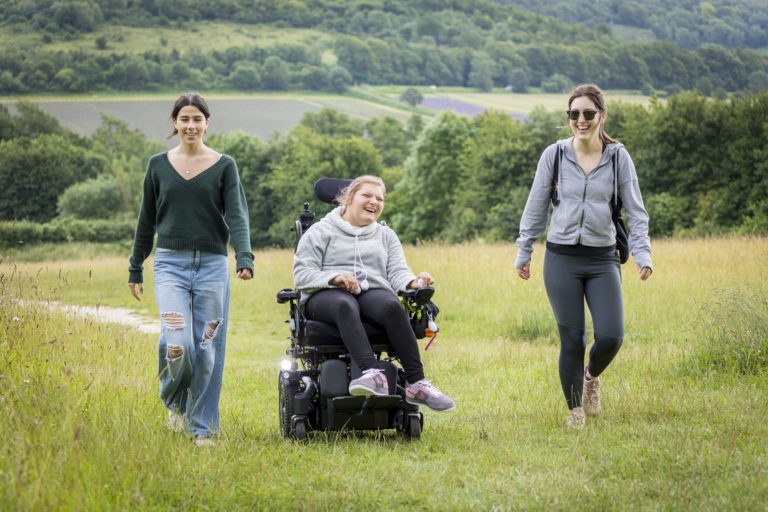 Three friends laugh as they walk across open grassland. Two are walking and one in an electric wheelchair.