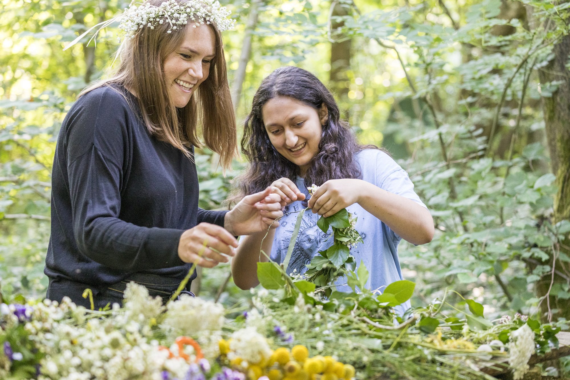 Two people in woodland, making a wreath from wild flowers. One has flowers in their hair.
