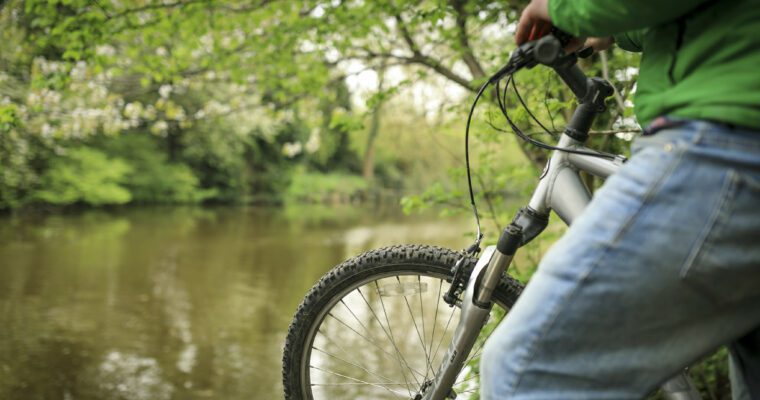 Close-up person on bike, stopped at river edge. River and surrounding trees in view.