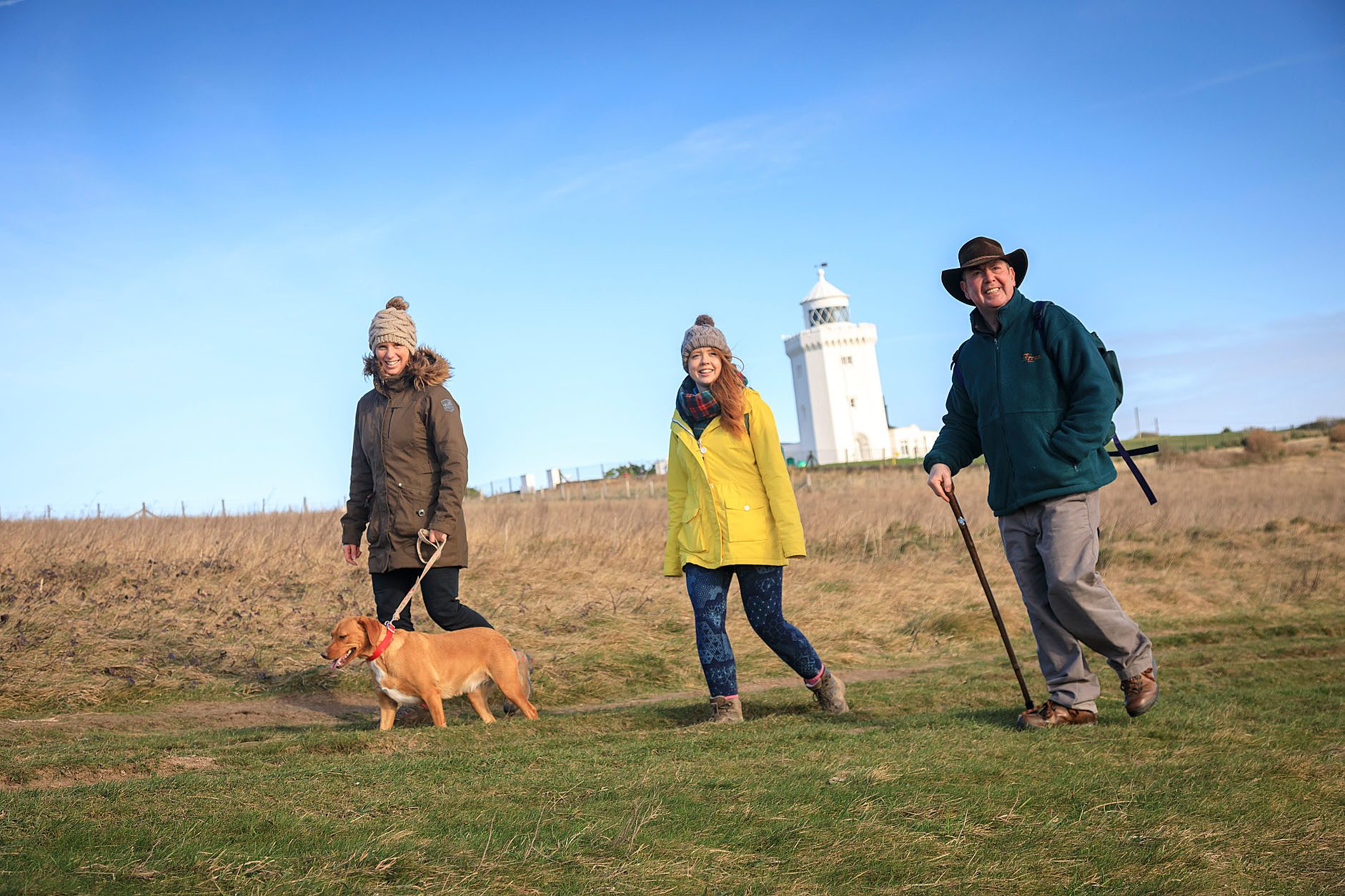 Three walkers with dog walking in front of white lighthouse