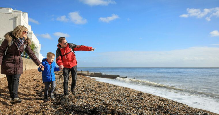 A family with a young child in winter coats walking along the shingle beach at St Margaret's Bay on a clear day