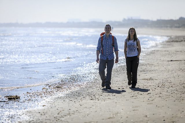 Two people walking on a sandy beach, towards camera. Sunny day, with calm sea on the left.