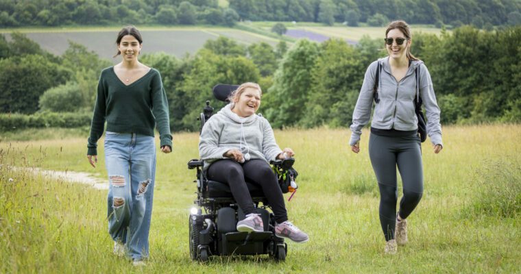 Three friends laugh as they walk across open grassland. Two are walking and one in an electric wheelchair.