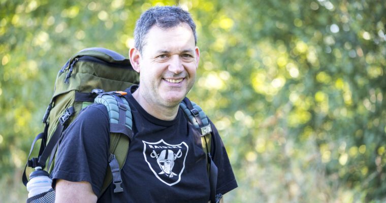 Philip Banfield smiling at camera, outdoors, with a rucksack on and greenery in background.