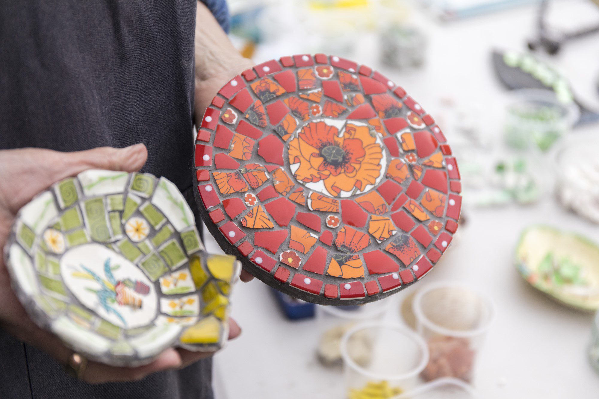Close-up person holding two handmade mosaic tiles. One is red and orange, the other white and green.