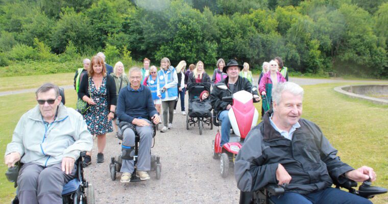 Group of walkers, some with mobility scooters and wheelchairs, on a wide path with trees in the distance.