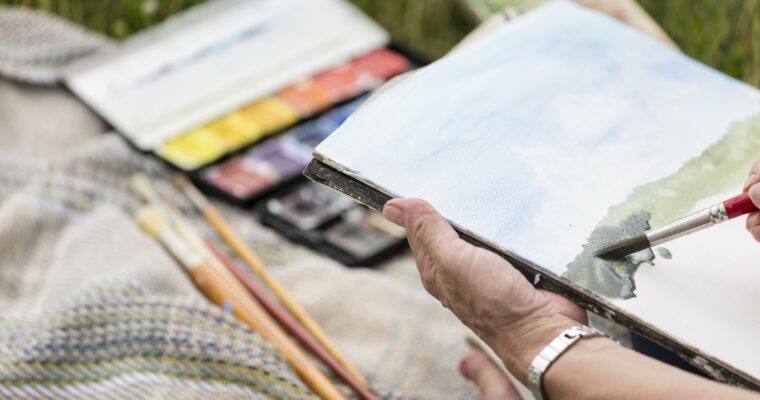 Close-up person holding a sketch pad, painting a picture in watercolours.