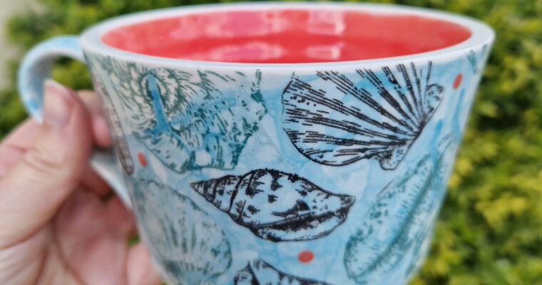 Close-up person holding a hand painted mug. The cup is blue with sea shells and red inside the cup.
