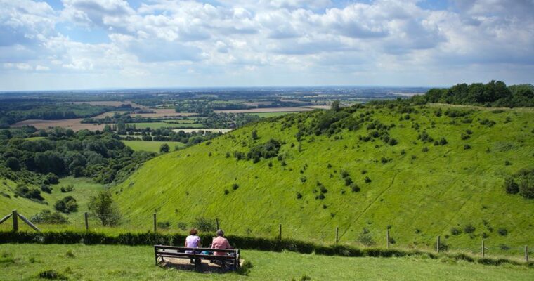 Two people sitting at bench, overlooking the view of Devil's Kneading Trough, on a sunny day.