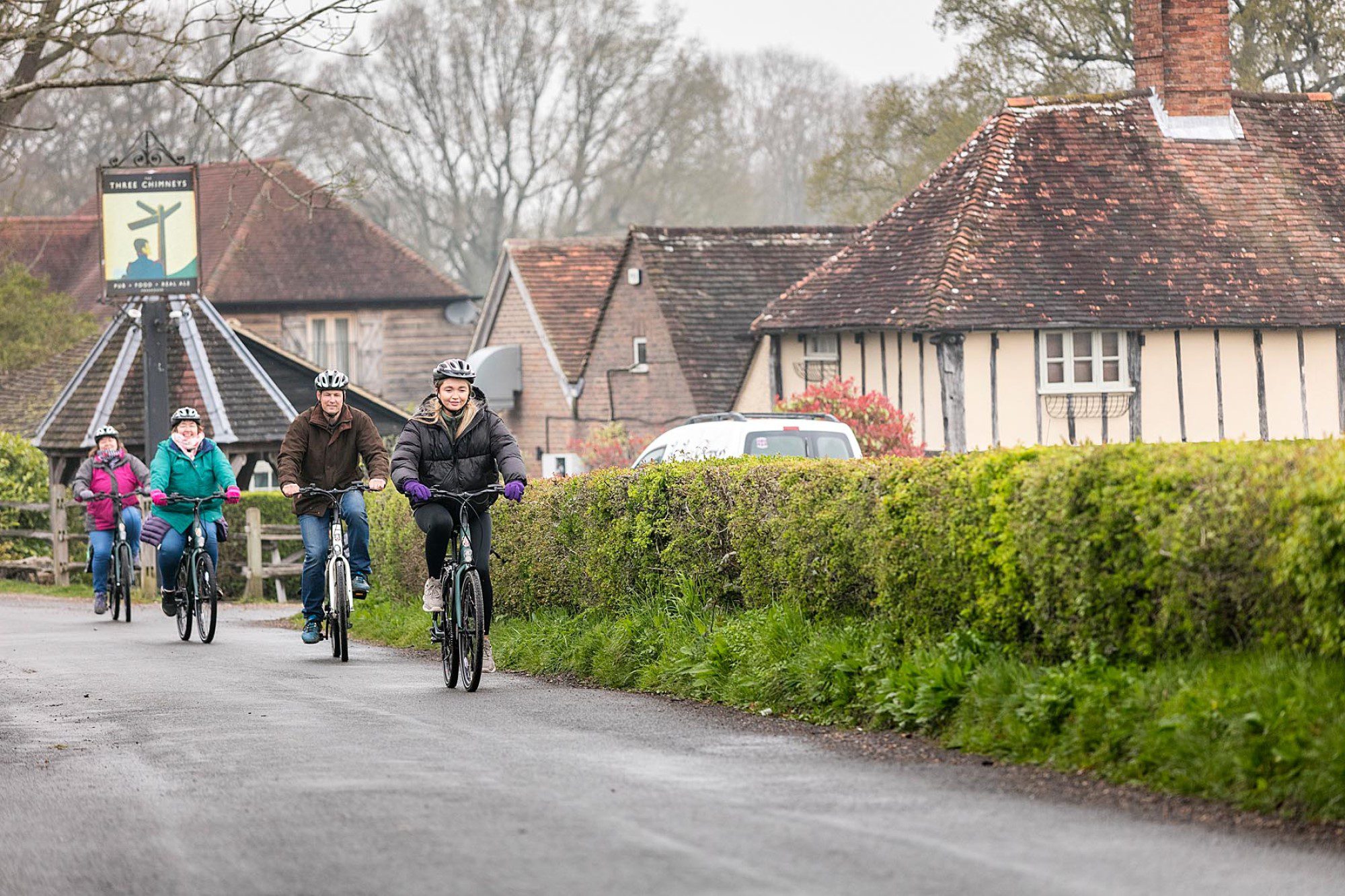 Four people cycling towards camera, on a road alongside the Three Chimneys pub.