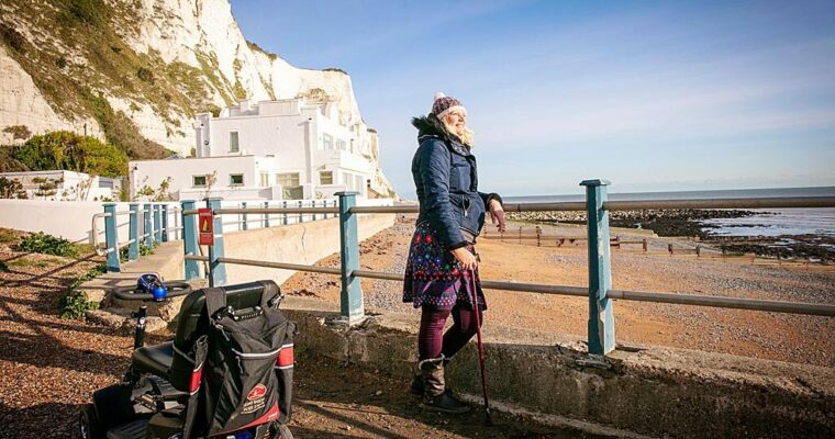 Person standing looking out over beach, mobility scooter nearby. Clear day with white cliffs in background.