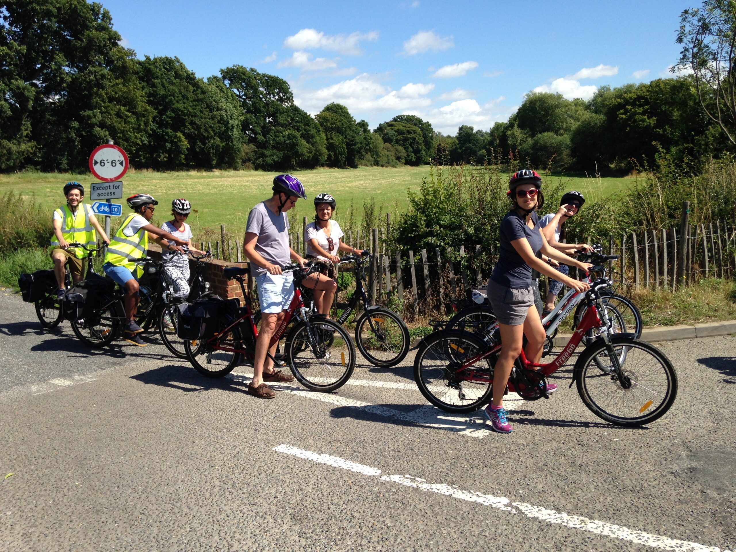 Group of people on bikes, stopped on a road, with grass fields and trees in background.