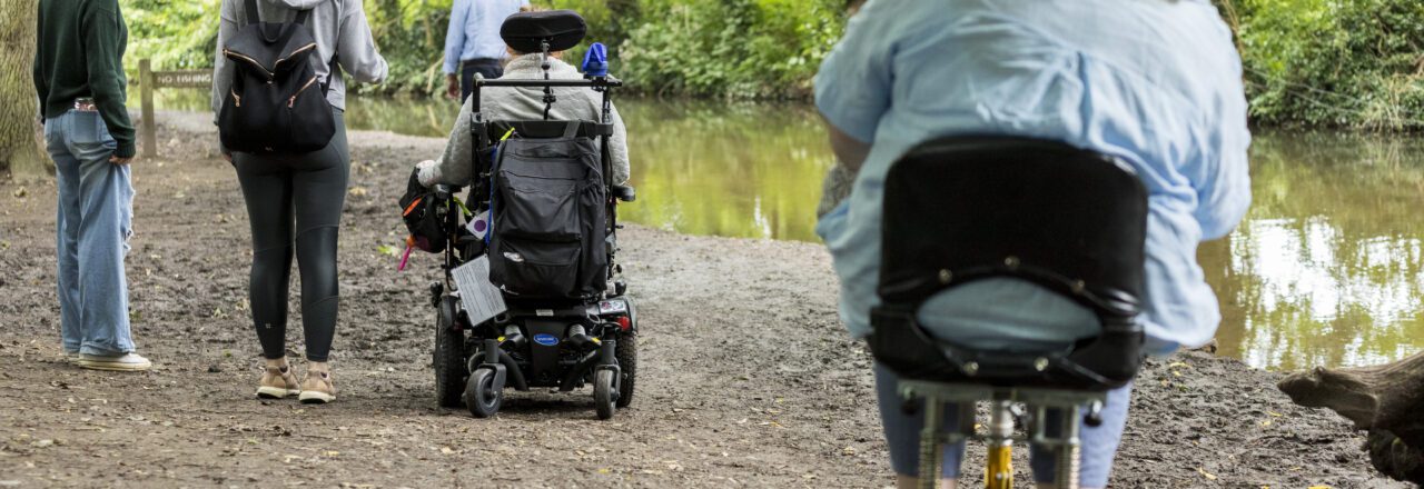 Group of walkers, two with mobility scooters, on a woodland path, near water and trees.