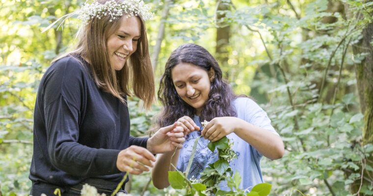 Two people in woodland, making a wreath from wild flowers. One has flowers in their hair.