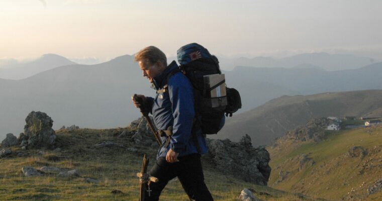Person with rucksack, walking on rockery and grass terrain. With hills in the distance.