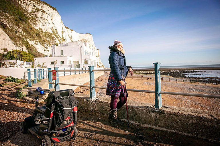 Person standing looking out over beach, mobility scooter nearby. Clear day with white cliffs in background.