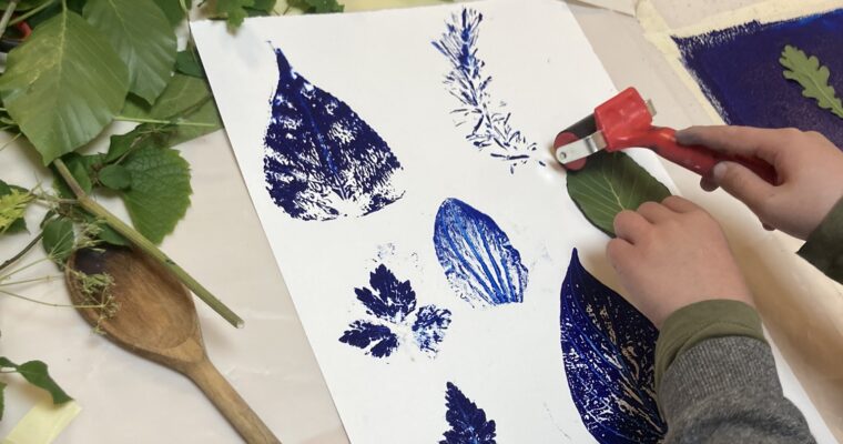 Close-up person's hands applying ink to a leaf, with other leaf ink images on paper.