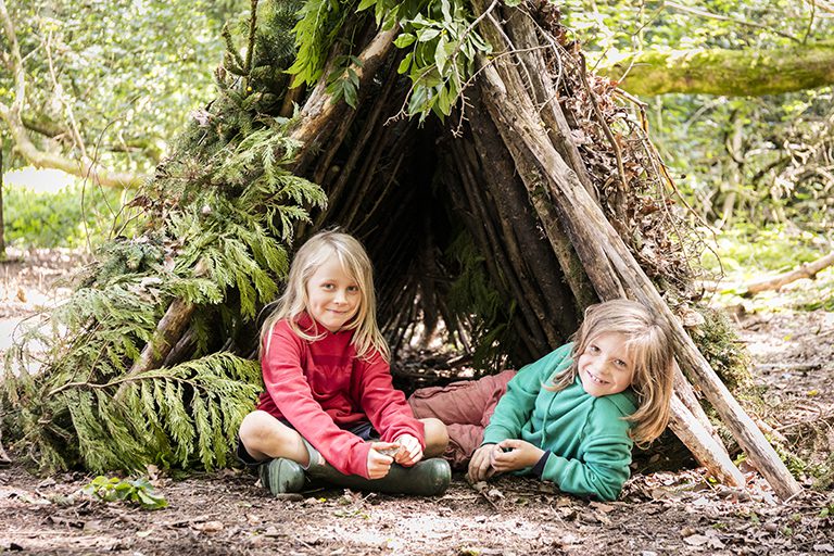 Two children sitting in the ground at the entrance to a wood built camp. Smiling and looking at camera.
