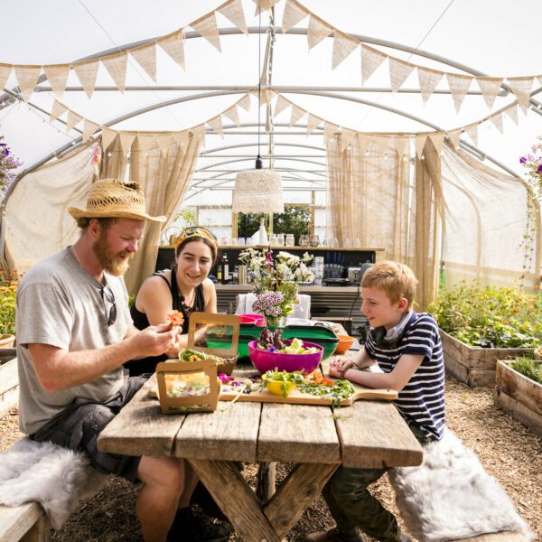 The Rebel Farmer and an adult & child sitting at a picnic style bench, in a polytunnel. Food is on the table and planter pots surround them.