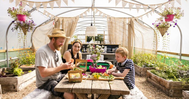 The Rebel Farmer and an adult & child sitting at a picnic style bench, in a polytunnel. Food is on the table and planter pots surround them.