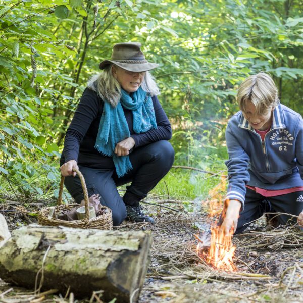 Adult and young adult kneeling on woodland floor, making a fire.