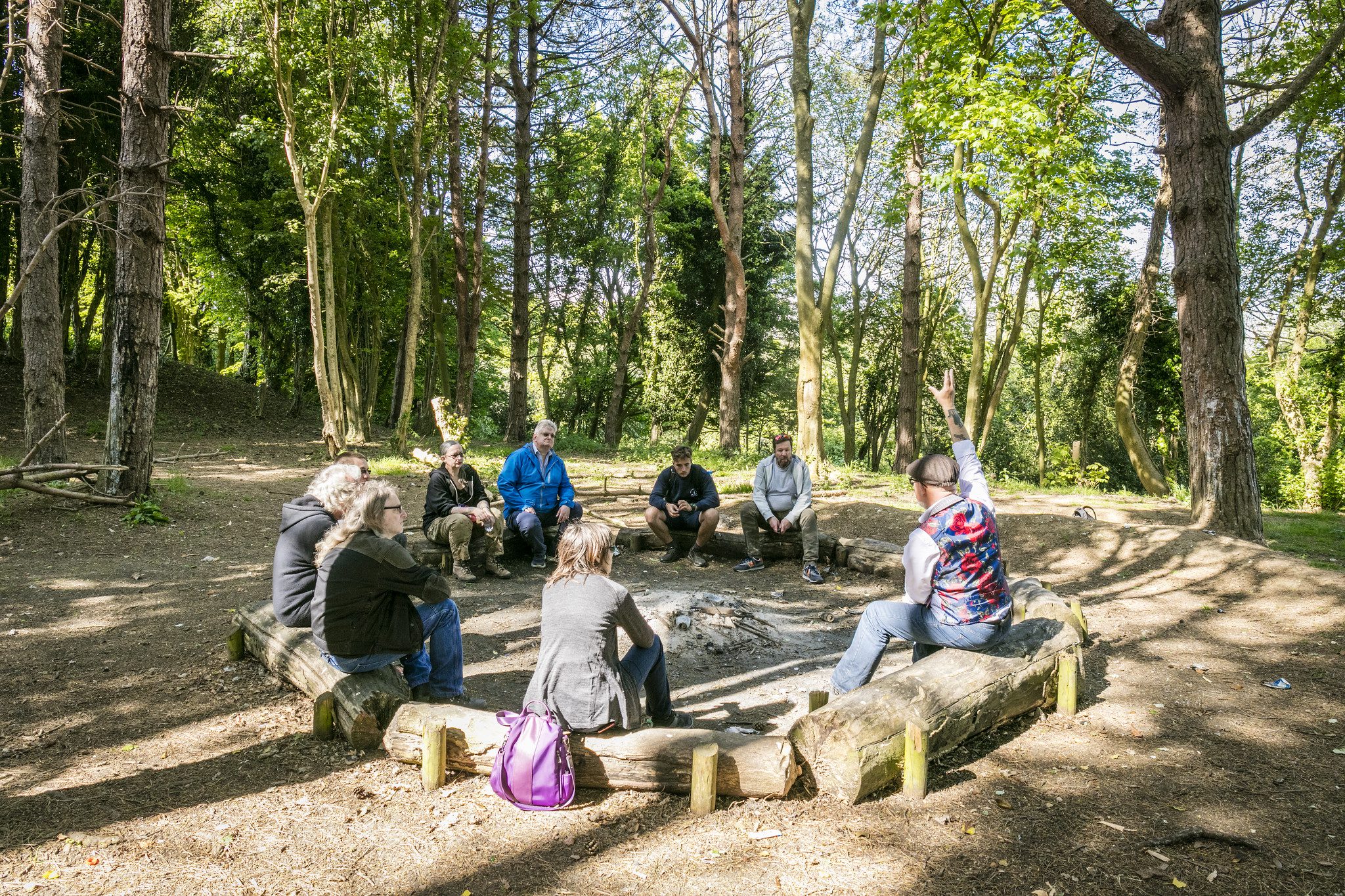 Group of people sitting in a circle in a woodland clearing. On a sunny day, one person has their hand raised.