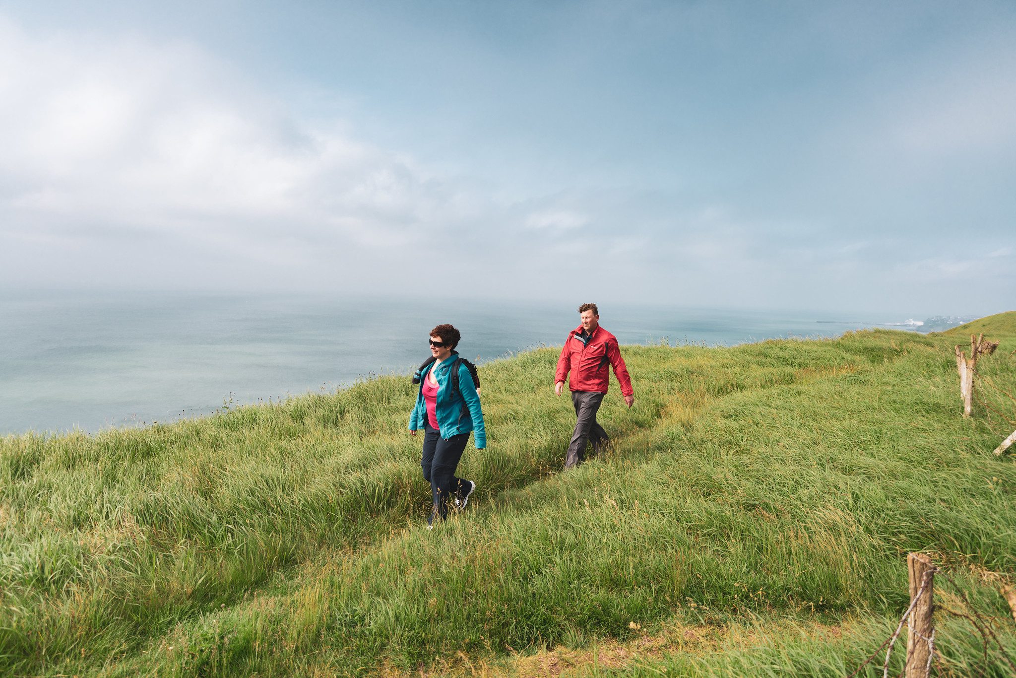 Two people walking on grass cliff top by the sea.