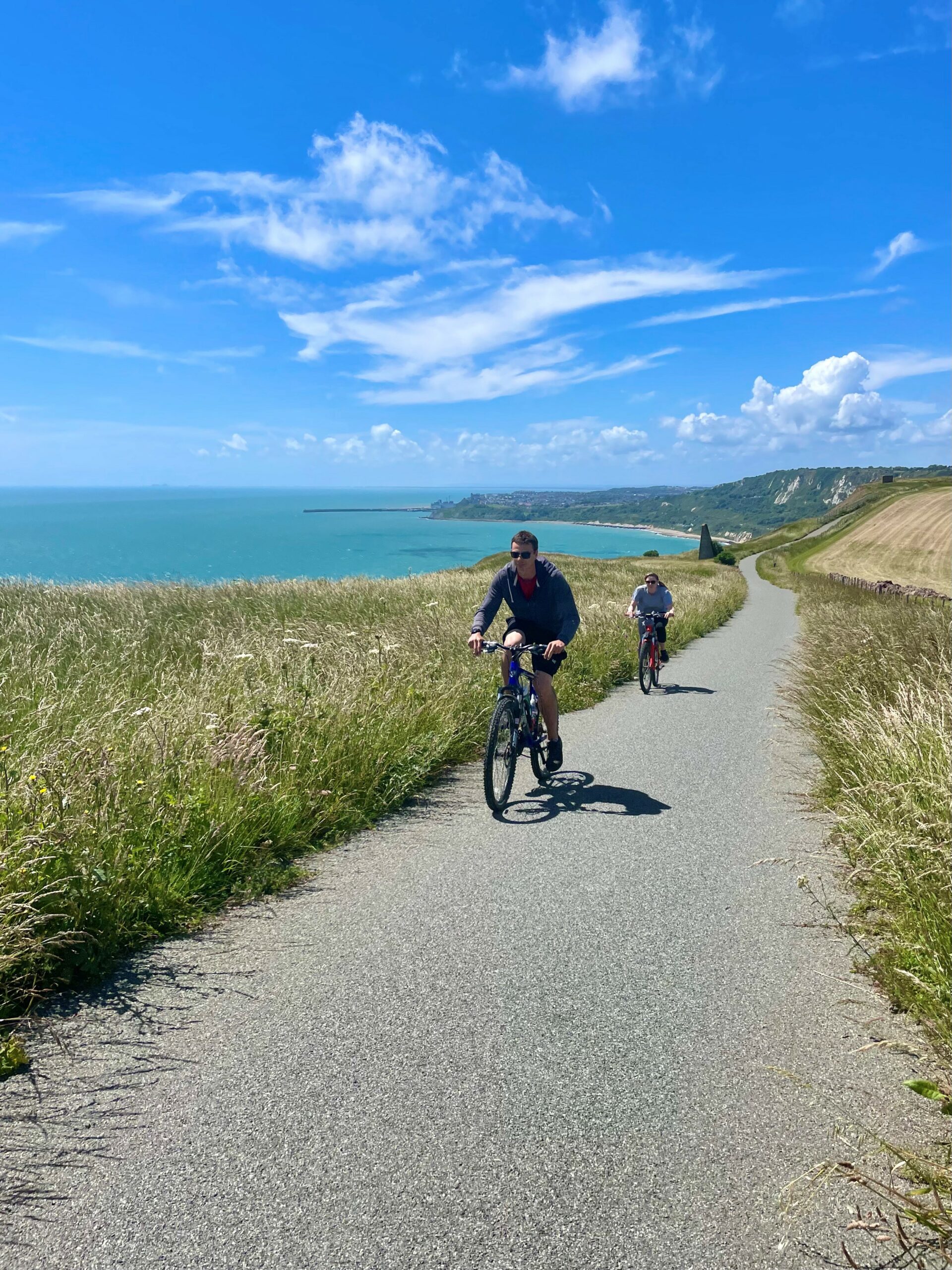 Two cyclists, on a cliff edge path, alongside the sea edge, on a sunny day.