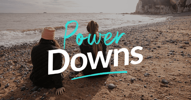 Two people sitting on beach, looking out to sea. Text Power Downs layered over image.