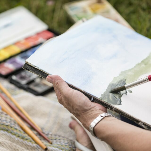 Close-up person holding a sketch pad, painting a picture in watercolours.