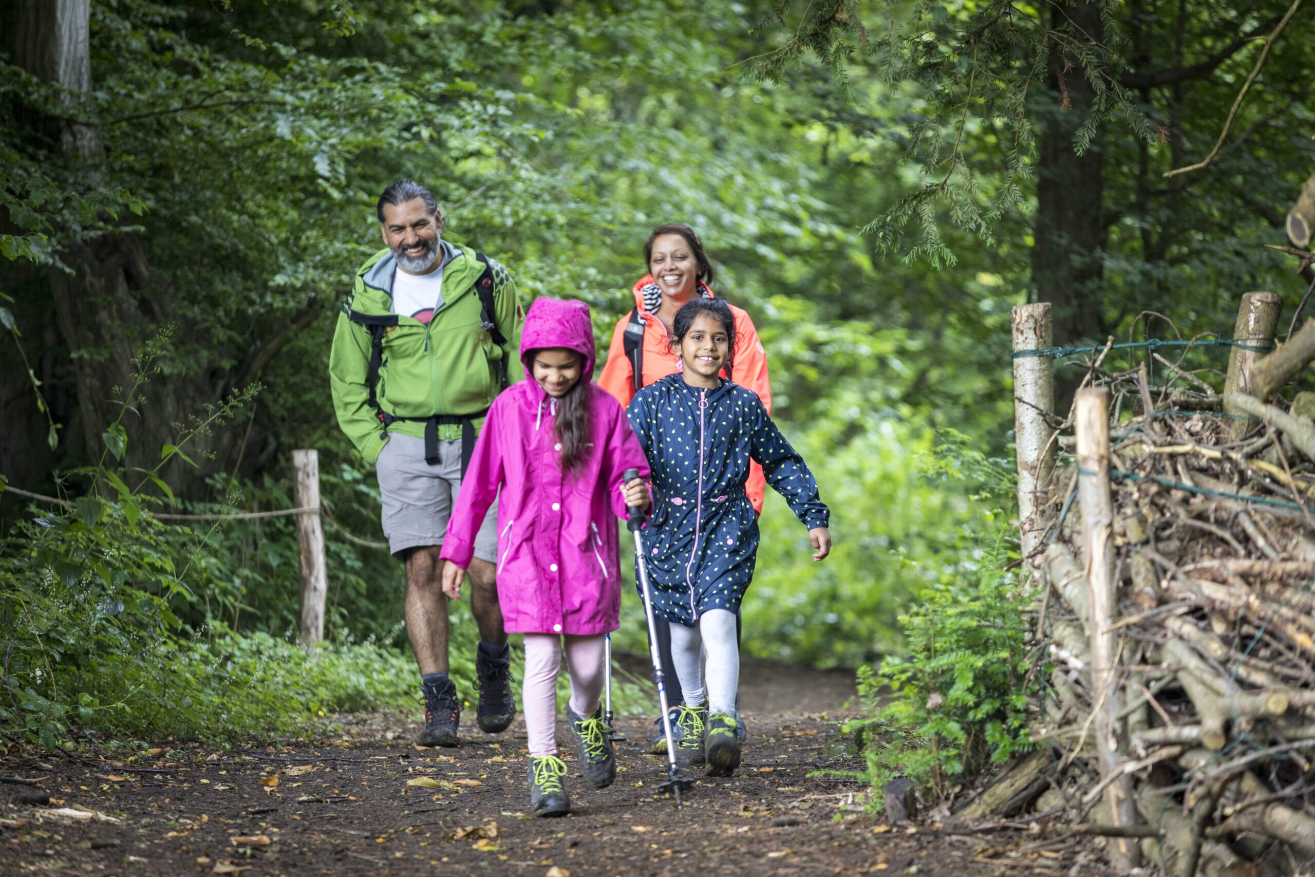 Two adults and two children walking towards camera, on a woodland path. Sunny day, everyone is smiling.