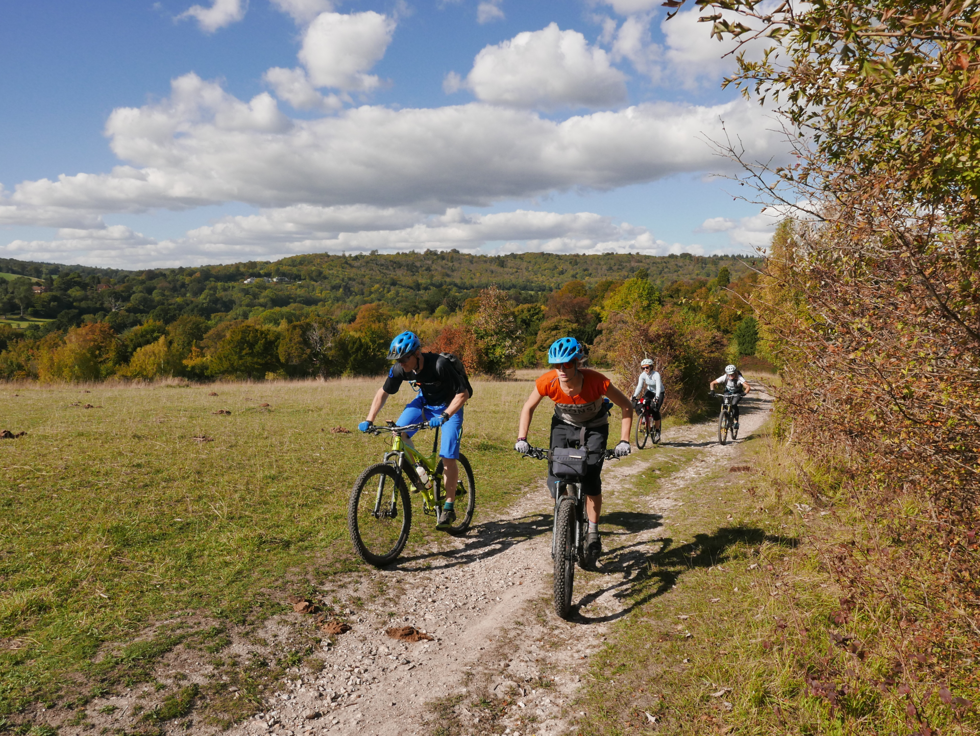 Group on cyclists on a dusty path, surrounded by grass fields and trees. Cycling towards the camera on a sunny day.