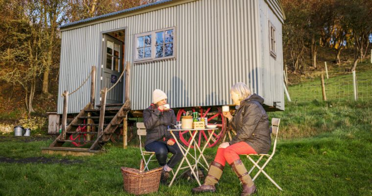 Shepherds hut in field, with trees in background. Two people seated at table and chair drinking and eating.