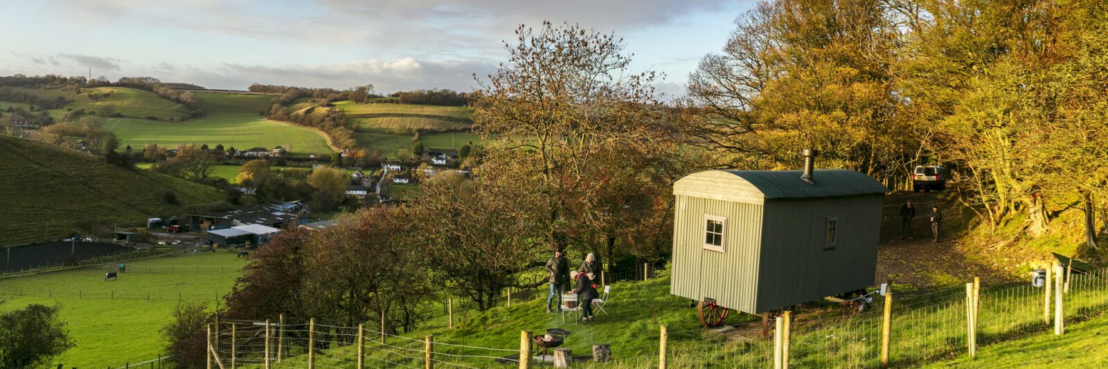 Shepherds hut and two people outside, with surrounding fields and trees.