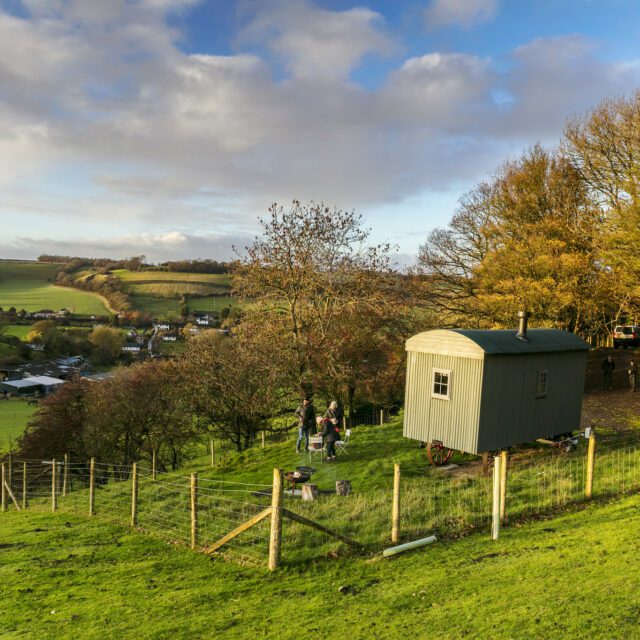 Shepherds hut and two people outside, with surrounding fields and trees.