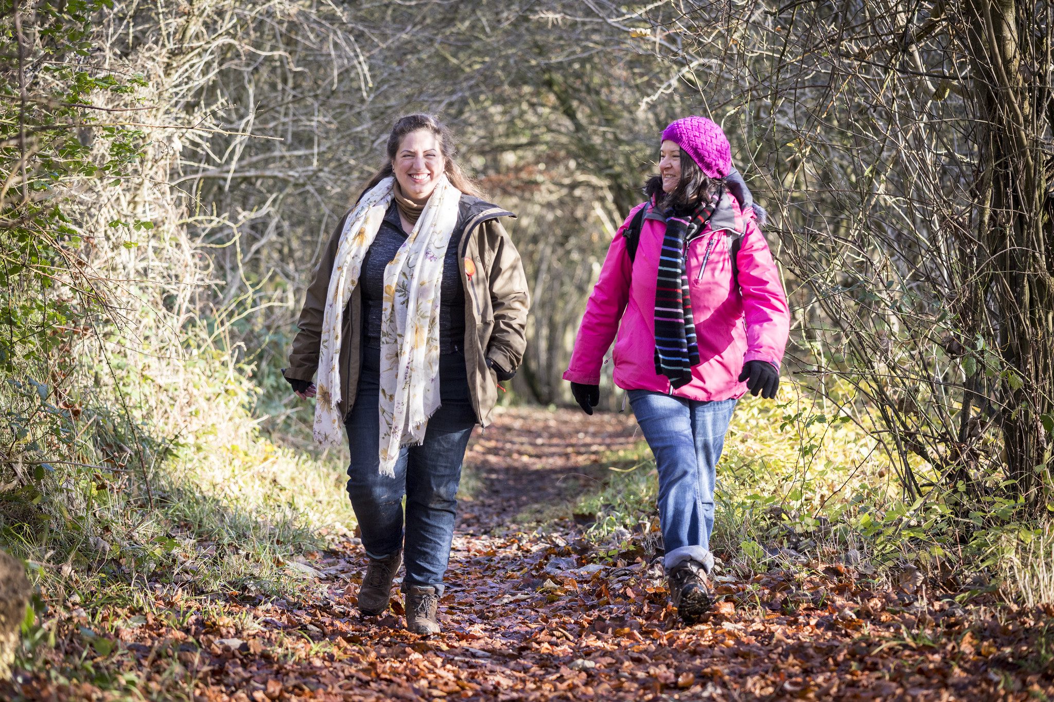 Two people walking towards camera, on woodland path with autumn leaves.