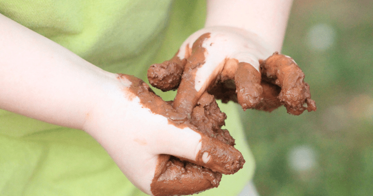 Child's hands playing with mud. The child has a green top on.
