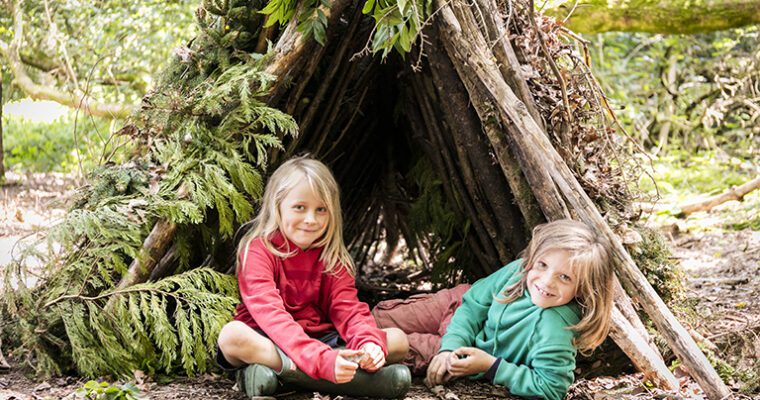 Two children sitting in the ground at the entrance to a wood built camp. Smiling and looking at camera.